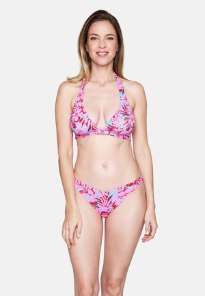 Frau steht, trägt einen pink-roten Blumen-Halfter-Bikini und lächelt vor einem schlichten hellen Hintergrund.