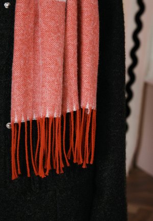 Tkaný šál v koralovej a bielej farbe, so štruktúrovaným vzorom a oranžovými třásňami, prehodený cez čierny odev so striebornými doplnkami.