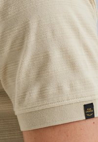 Beige kortærmet skjorte med strukturerede vandrette striber, ribbet manchet og et lille sort mærke med hvid tekst ved kanten.