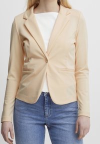 Blazer v broskvové barvě vyrobený z měkké tkaniny, s jedním knoflíkem, klopami a dvěma bočními kapsami, nošený přes bílý top.