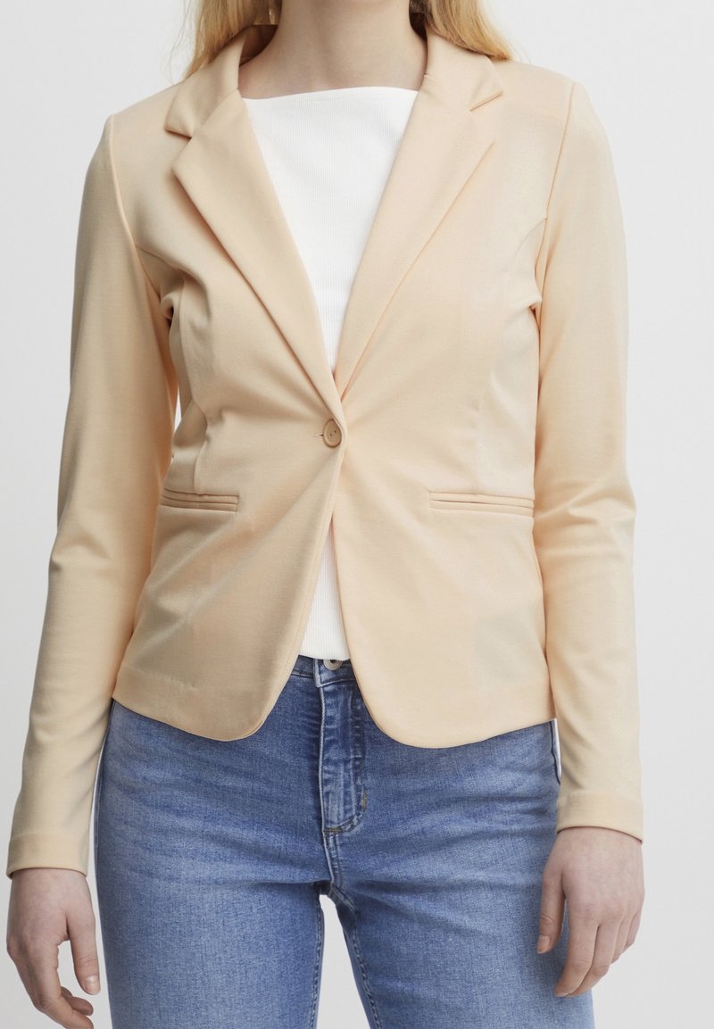 Blazer v broskvové barvě vyrobený z měkké tkaniny, s jedním knoflíkem, klopami a dvěma bočními kapsami, nošený přes bílý top.