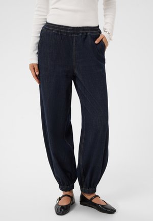 Personne portant un pantalon en jean bleu foncé à taille élastique avec chevilles froncées et des chaussures plates Mary Jane noires.