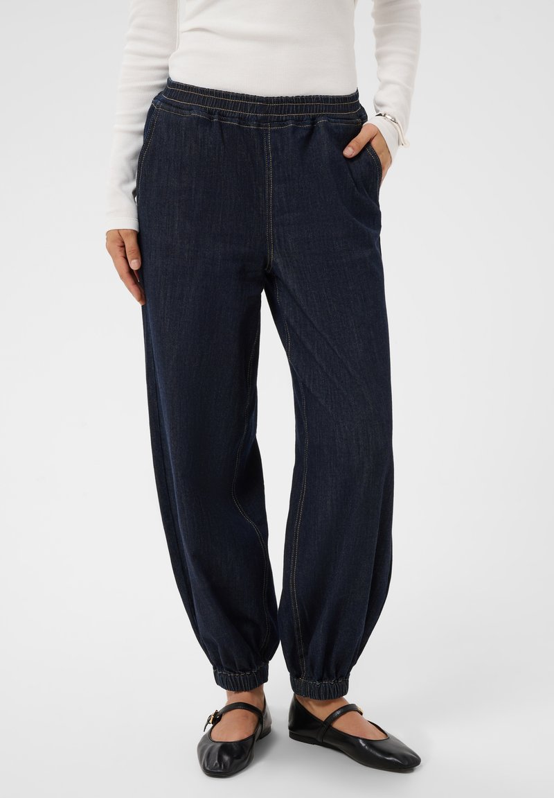 Personne portant un pantalon en jean bleu foncé à taille élastique avec chevilles froncées et des chaussures plates Mary Jane noires.