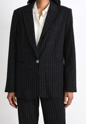 Vrouw gekleed in een zwart krijtstreepblazer met bijpassende broek en een wit overhemd met knopen, staand tegen een effen achtergrond.