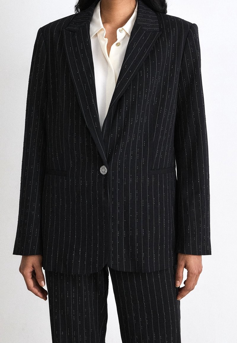 Femme portant un blazer noir à fines rayures assorti à un pantalon et une chemise blanche boutonnée, debout devant un fond uni.