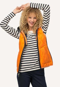 Gilet trapuntato arancione con rifiniture blu navy sopra una maglietta a righe bianca e blu navy. Il gilet presenta una chiusura a zip e tasche frontali.