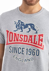 Grå bomuld t-shirt med et blåt løvegrafik og fed rød tekst med mærket "Lonsdale" og "Siden 1960" med cremefarvede accenter.