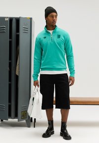 Τουρκουάζ φούτερ με φερμουάρ και λογότυπο, μαύρα σορτς μέχρι το γόνατο και μαύρα δερμάτινα μποτάκια μέχρι τον αστράγαλο. Λευκή τσάντα με πράσινες λεπτομέρειες. Ατσάλινα ερμάρια στο φόντο.