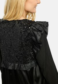 Bluză neagră cu textură paietată pe spate și accente volanate, material neted și siluetă structurată.