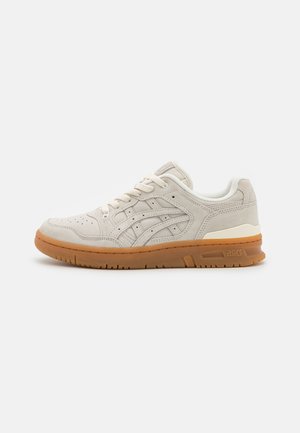 Zapatilla deportiva de gamuza beige claro con perforaciones, cordones a juego y suela de goma color caramelo, mostrada de lado sobre un fondo blanco.