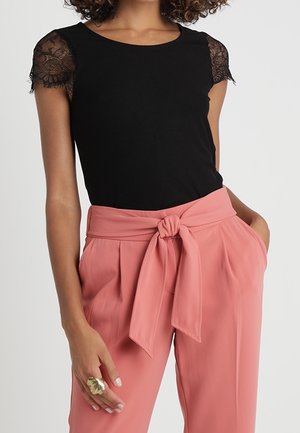 Femme portant un haut noir avec des manches courtes en dentelle et un pantalon rose taille haute, noué à la taille avec une large ceinture en tissu.