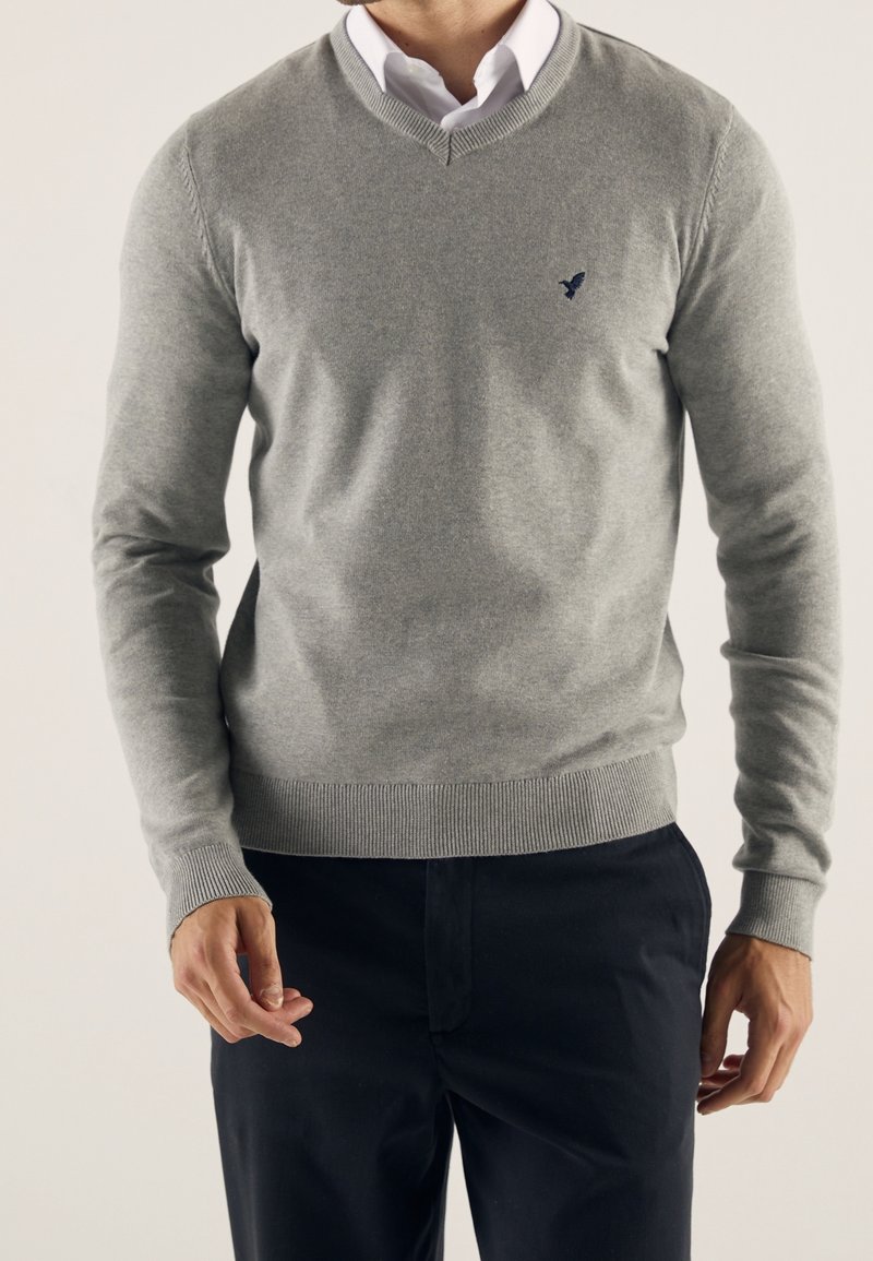 Homme portant un pull gris en V sur une chemise blanche à col et un pantalon foncé, debout avec une posture détendue.