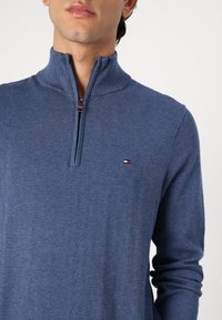 Blå zip-tröja i mjukt stickat tyg, med ståkrage och en liten logotyp på vänster bröst.