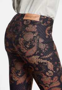 Jeans negros con un vibrante patrón de paisley en tonos rojos y dorados, con una etiqueta de marca de cuero beige en la cinturilla trasera y los bolsillos traseros.