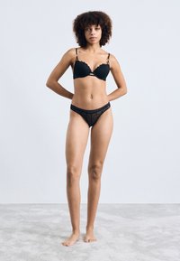 Sutien din dantelă neagră cu margini ondulate și bretele subțiri, asortat cu un thong negru transparent, cu modele geometrice. Textură netedă.