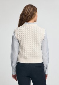 Cremfarbener Kabelstrick-Weste mit geripptem Saum und ärmellosem Design, getragen über einem blau-weißen gestreiften Hemd mit klassischem Kragen.