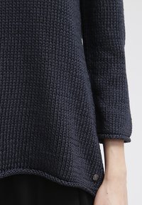 Pull en maille épaisse bleu marine avec manches longues et ourlet arrondi porté sur le torse d'une personne, bras visible.