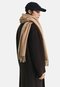 Vrouw die naar rechts kijkt, met een zwarte baseballcap, een donkerbruine jas en een grote camel-kleurige sjaal over haar schouders gedrapeerd.