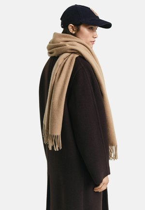 Vrouw die naar rechts kijkt, met een zwarte baseballcap, een donkerbruine jas en een grote camel-kleurige sjaal over haar schouders gedrapeerd.