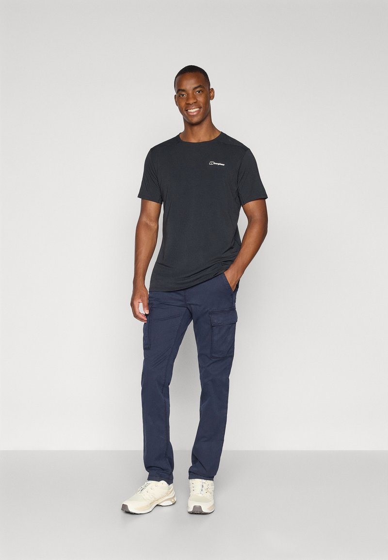 Un homme souriant se tient avec une main dans sa poche, portant un t-shirt noir, un pantalon cargo bleu marine et des baskets blanches sur un fond clair et uni.