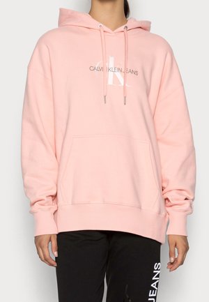 Osoba w jasnoróżowej bluzie z kapturem Calvin Klein Jeans ze sznurkami i czarnych spodniach z białym napisem na lewej nodze.