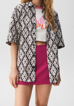 Camisa de manga corta de patrón en blanco y negro, llevada abierta sobre un top corto blanco con texto naranja, combinada con una falda mini ajustada rosa.