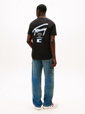 T-shirt preta de algodão com texto gráfico azul claro nas costas, combinado com jeans de ganga azuis e ténis brancos com detalhes em vermelho e azul.