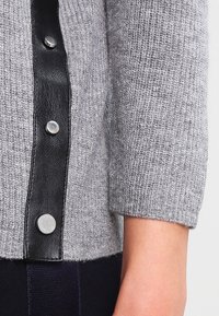 Gros plan d'une manche de pull gris tricoté avec une patte de boutonnage en cuir noir et des boutons-pression argentés sur un bras.