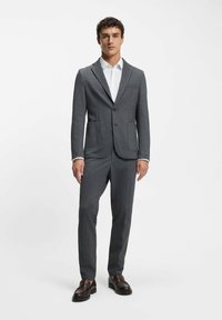 Man draagt een nauwsluitend grijs pak met een wit overhemd en bruine leren loafers, staand tegen een effen witte achtergrond.