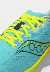 Φωτεινό τιρκουάζ αθλητικό παπούτσι με φλούο κίτρινες κορδόνια, μαύρο κλείδωμα κορδονιών και μαύρο-κίτρινο λογότυπο στο πλάι.