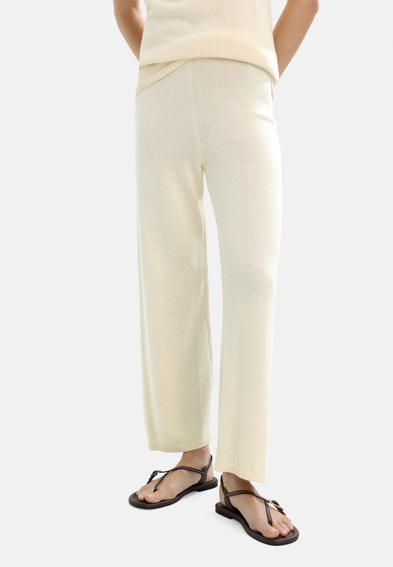 Jas beige'owe dzianinowe spodnie o szerokich nogawkach, teksturowanej powierzchni i ściągaczu w prążki. W połączeniu z brązowymi płaskimi sandałami, prezentującymi otwarty czubek.