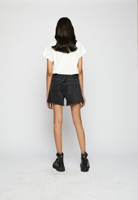 Pantalones cortos de mezclilla negra con un dobladillo deshilachado, combinados con una camiseta blanca de manga corta y botas negras hasta el tobillo. Vista desde la parte trasera.