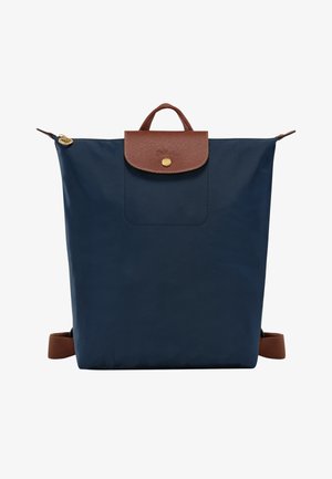Navy blue backpack with a textured brown leather flap, gold zipper, and front pocket. Adjustable brown straps for carrying.