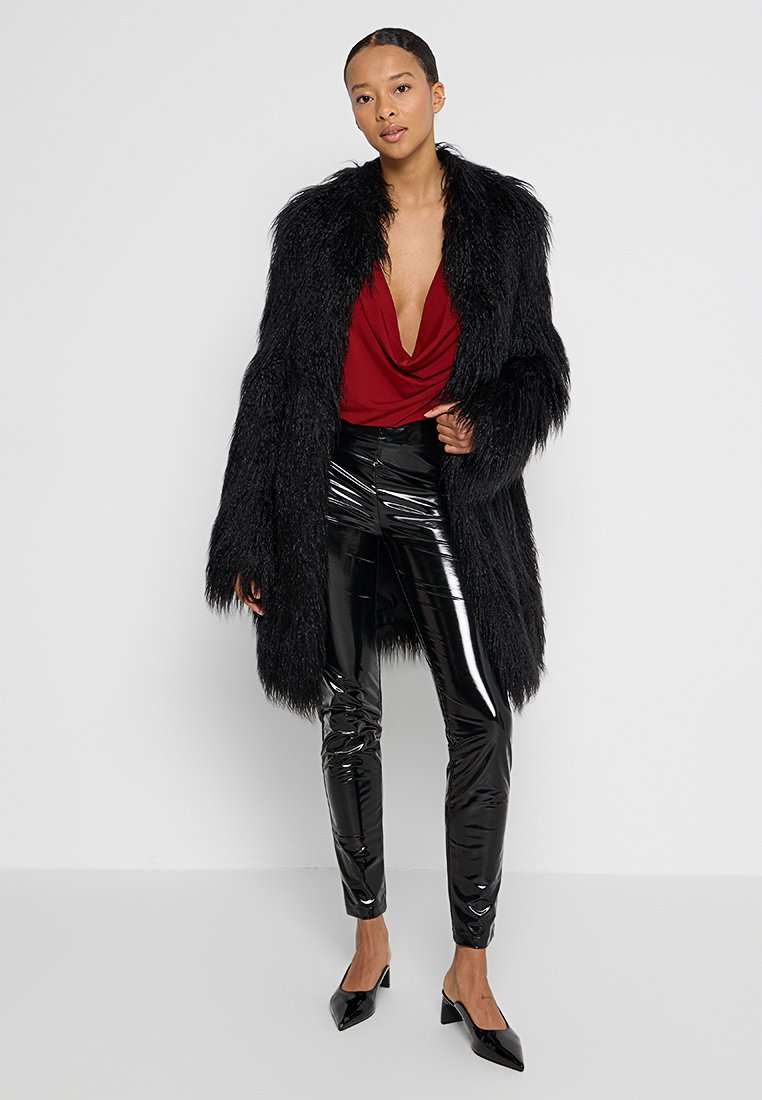 Frau in einem schwarzen Kunstfellmantel, rotem tiefen V-Ausschnitt-Shirt, glänzenden schwarzen Hosen und schwarzen spitzen High Heels, die vor einem weißen Hintergrund steht.