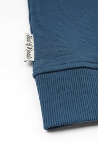 Sininen resorikangas, jossa on käännetty hihansuu ja valkoinen, mustalla tekstillä kudottu etiketti. Pinta on teksturoitu, yksinkertainen muotoilu.