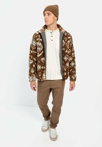 Bruine fleecejack met een patroon en witte en grijze ontwerpen, gedragen over een witte henleyshirt, gecombineerd met een paar bruine broeken en witte sneakers.