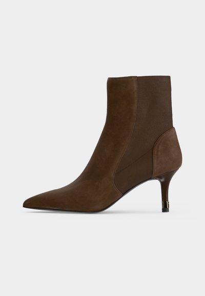 Bottine en daim marron avec bout pointu et talon haut et fin avec un accent doré sur fond neutre.