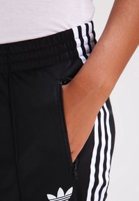 Svarta sportbyxor med vita ränder, en ficka med dragkedja på sidan och en vit logotyp på vänster sida av midjebandet.