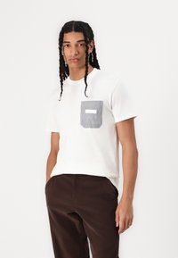 Tricou alb din bumbac cu mâneci scurte, având un buzunar cu dungi în albastru marin și alb. Pantaloni din catifea corb cu un ton de maro completează ținuta.