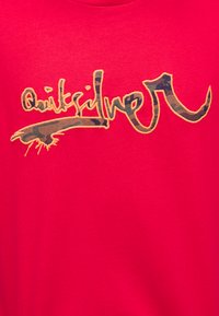 Φωτεινό κόκκινο ύφασμα με κείμενο "Quiksilver" σε καφέ καμουφλάζ μοτίβο με πορτοκαλί περίγραμμα κεντήματος.