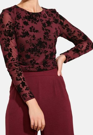 Kvinde iført en bordeaux langærmet top med sorte blomsterprint og matchende bukser med høj talje, hånd hvilende på hoften.