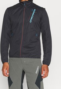 Schwarze, leichte Zip-Jacke mit blauen Akzenten und hohem Kragen. Verfügt über zwei Seitentaschen und ein Logo entlang der Vorderseite.