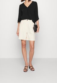Schwarzes, langärmliges V-Ausschnitt-Oberteil, lockere Passform, kombiniert mit beigen Shorts mit Falten. Das Model hält eine kleine schwarze Tasche und trägt schwarze Sandaletten mit Absatz.