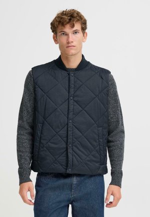 Marineblå quiltet væ vest med trykknapper og ribbet krave, båret over en grå striklangærmet skjorte. Klassisk pasform, funktionelt og varmt design.