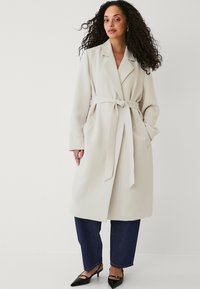 Lyse beige indpakningsfrakke med bælte, brede kraver og sidelommer. Båret sammen med mørkeblå jeans og spidse sorte hæle.