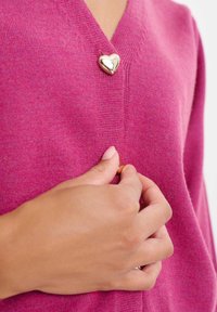Różowy kardigan dziany z dekoltem w serek i złotym guzikiem w kształcie serca przy dekolcie. Miękka tekstura, dopasowany fason, ukazujący ułożenie dłoni.