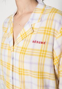 Rutig skjorta med knappar i gult, vitt och lavendel. Mjukt strukturerat tyg med krage och röd broderad detalj med texten "RÉSUMÉ".