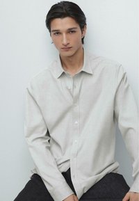 Camicia grigia chiara con bottoni realizzata in tessuto strutturato, con colletto, maniche lunghe e sottili dettagli di cucitura, abbinata a pantaloni scuri.