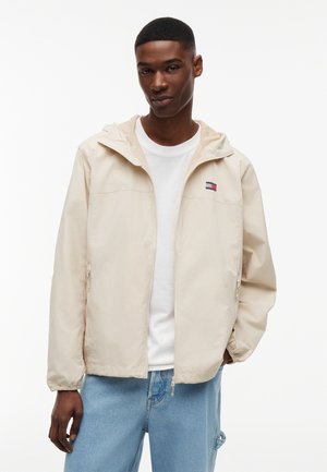 Νέος άνδρας που φοράει μπεζ μπουφάν με κουκούλα πάνω από λευκό πουκάμισο και ανοιχτόχρωμα τζιν, στέκεται μπροστά σε απλό λευκό φόντο.