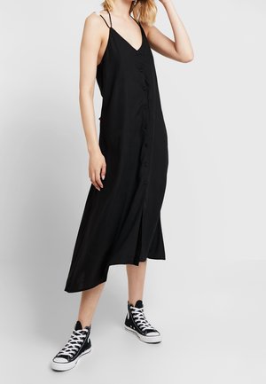 Schwarz midi Kleid mit V-Ausschnitt, Knopfleiste und seitlicher Schlitze, aus leichtem Stoff. Kombiniert mit schwarzen High-Top Sneakers.